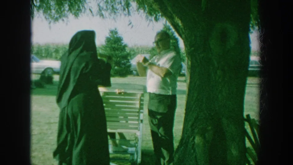 1964 New York home-movie frame showing a dark cloaked figure near a wooden bench and a man in glasses holding corn under a large tree.