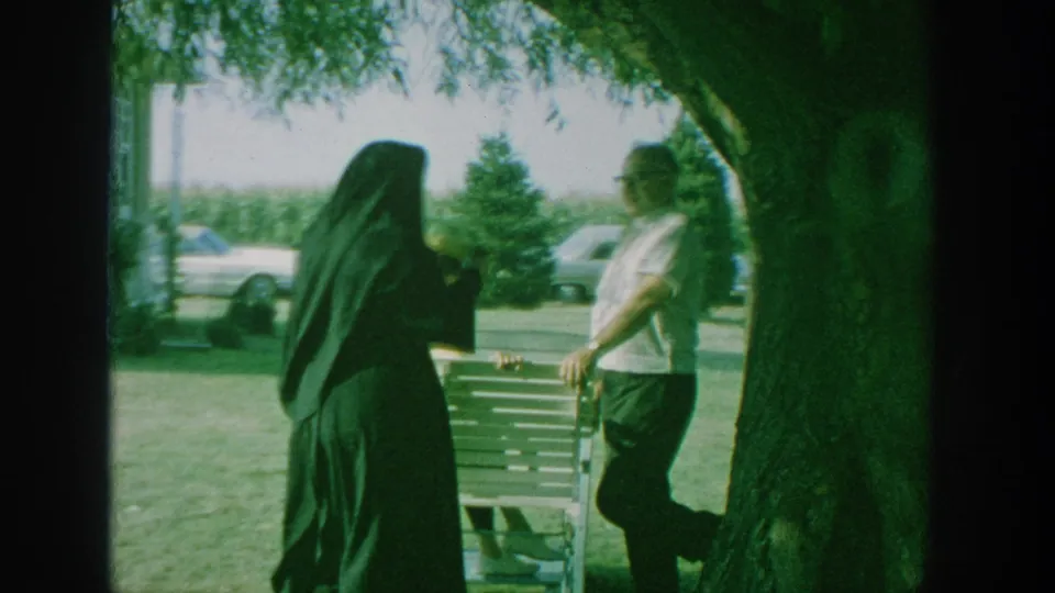 1964 New York home-movie frame with a dark cloaked figure and a man standing beside a wooden bench under a large tree, with cars beyond the lawn.