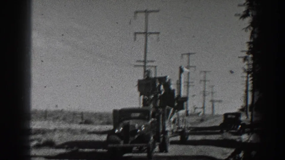 1936 California 8mm frame of a classic car towing a loaded flatbed trailer past utility poles on an open rural road.