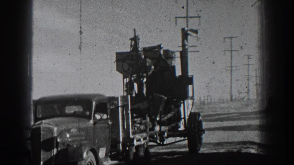 The trailer and towing car pass the camera, showing stacked cargo, a riding man, road shoulder, and utility poles in 1936 California footage.