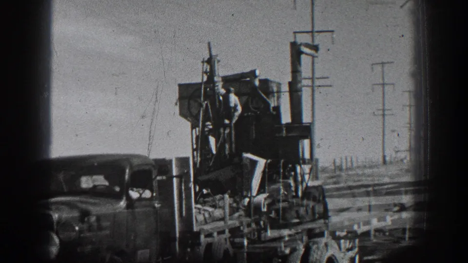 1936 California home-movie frame showing a worker near machinery mounted on a classic truck along a roadside with utility poles.