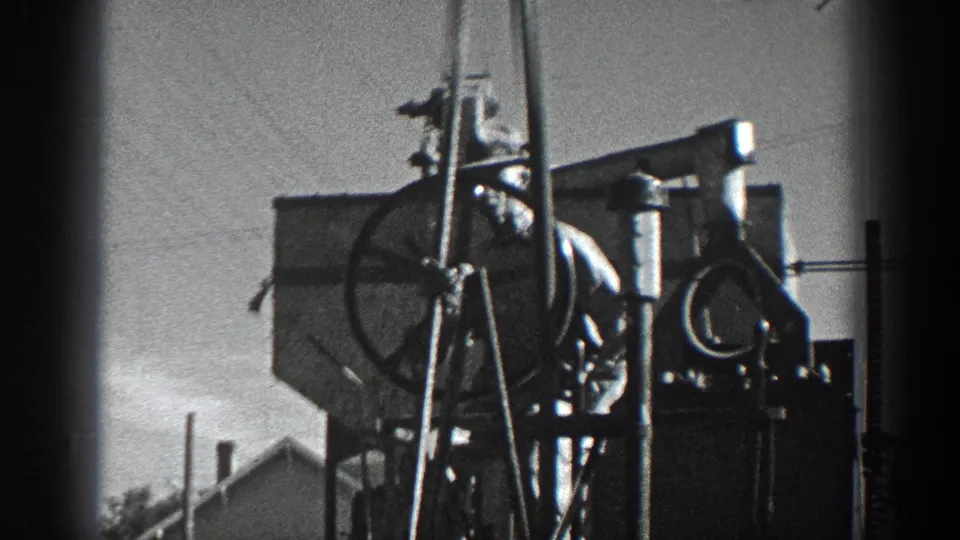 Close handheld 8mm view of wheel-operated machinery with a partly visible person and utility infrastructure in California, 1936.
