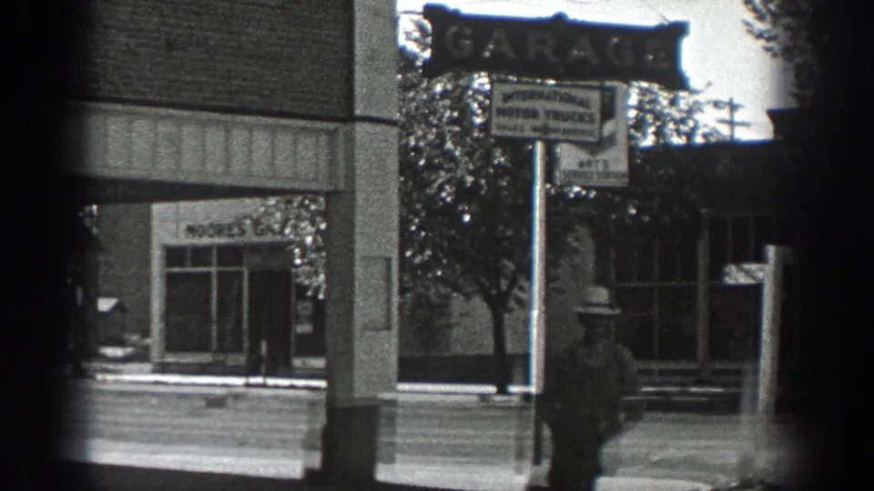 California town street corner in 1936 with a visible garage sign, storefronts, utility pole, and pedestrian in period attire.