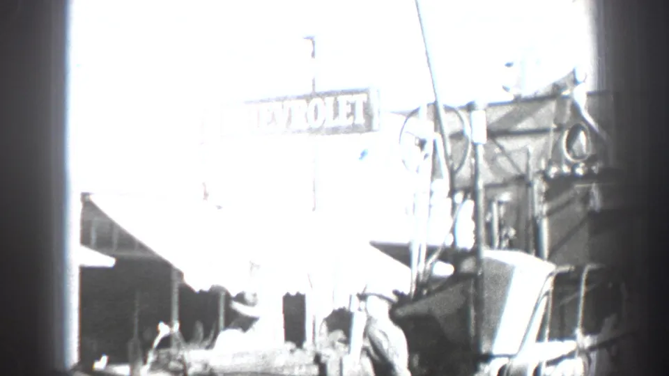 Black-and-white 1936 California home-movie frame showing a bright street scene with Chevrolet signage, period automobiles, and people near a commercial garage or dealership area.