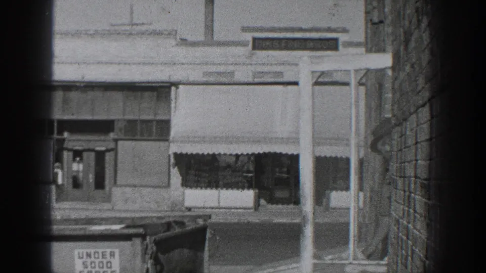 Black-and-white 1936 California street frame showing storefront awnings across the road, a signpost, a brick wall, and part of a period car in the foreground.