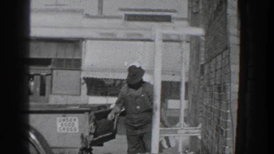 Black-and-white 1936 California home-movie frame showing a man in a hat beside a period car near a brick wall, storefront windows, and a sidewalk sign advertising used good cars.