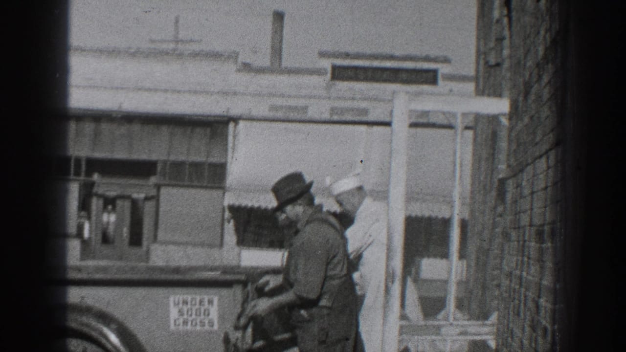A man in overalls handles cargo at a truck while another man in a white cap stands behind him.