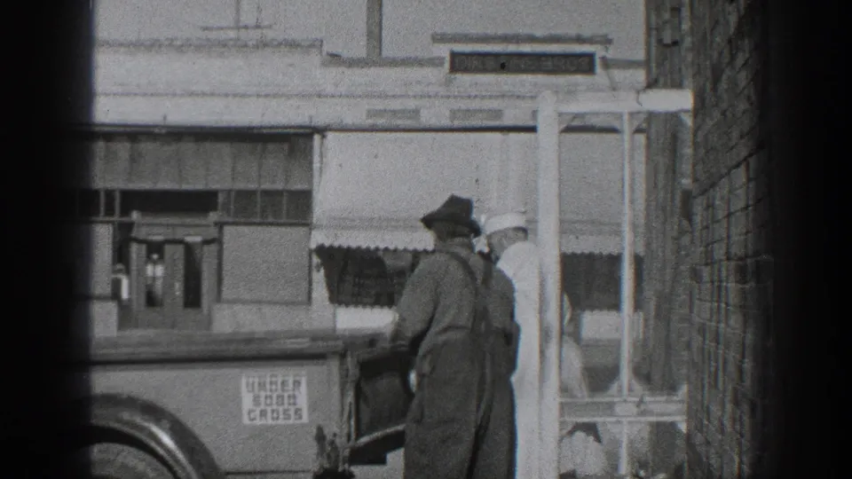 1936 California 8mm frame showing two adult men at a truck in a narrow alley, with commercial windows and an awning across the street.