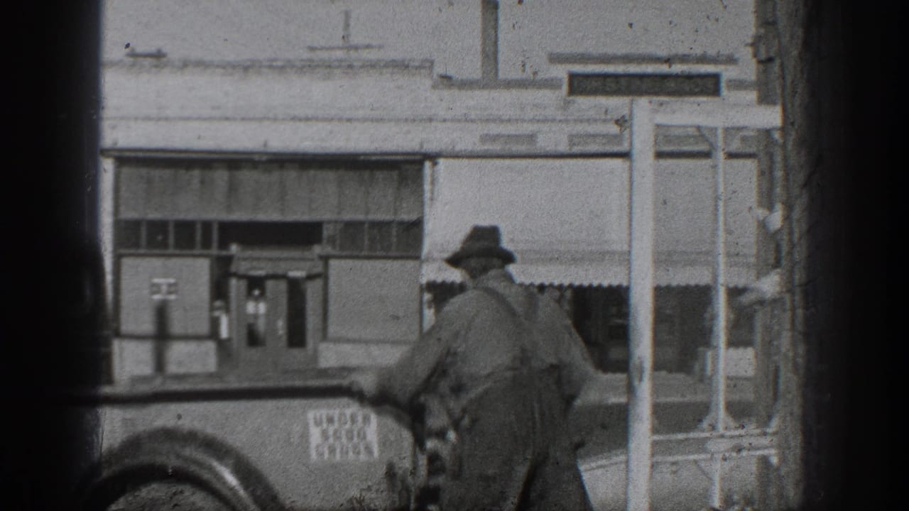 1936 California home-movie frame showing a worker in hat and overalls beside an older truck outside industrial buildings.