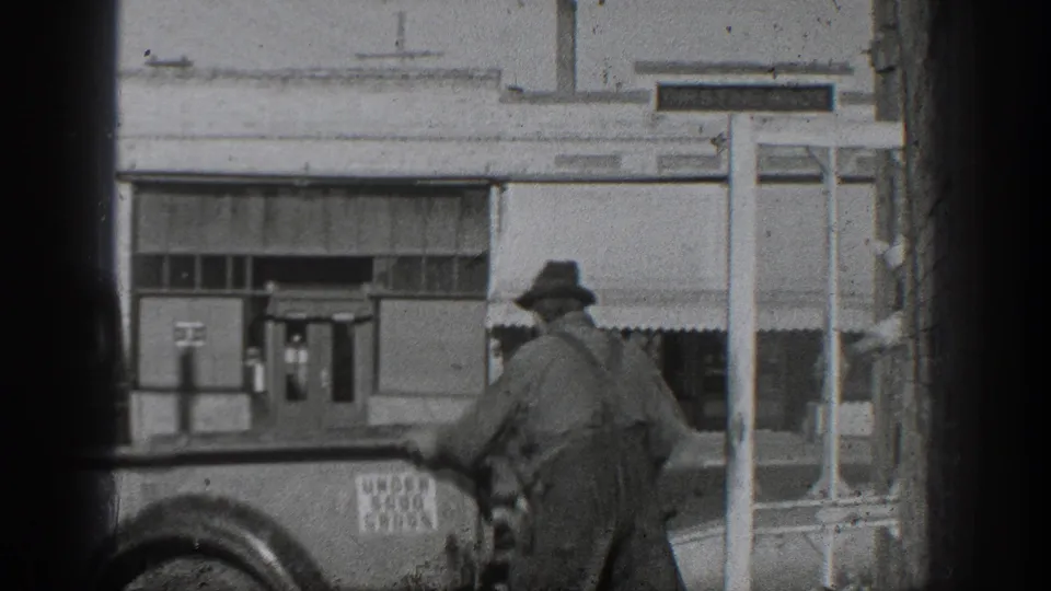 1936 California home-movie frame showing a worker in hat and overalls beside an older truck outside industrial buildings.