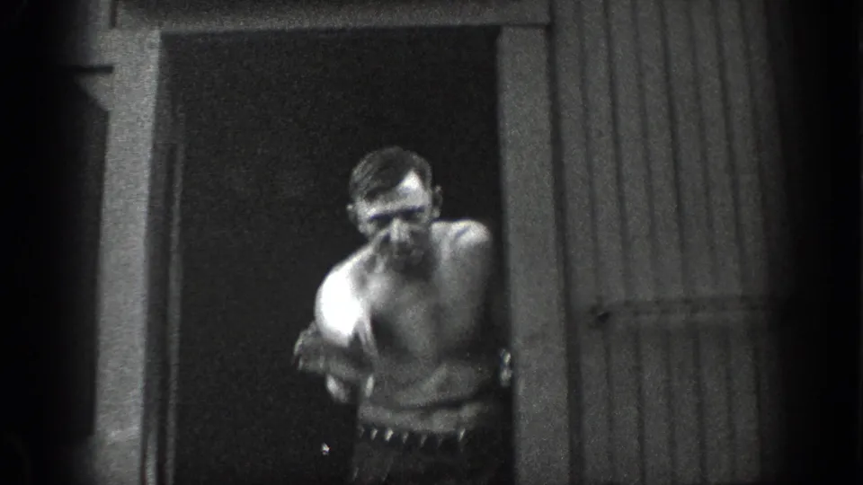 1936 California home-movie frame of a shirtless man standing in or near a doorway beside a sided wall.