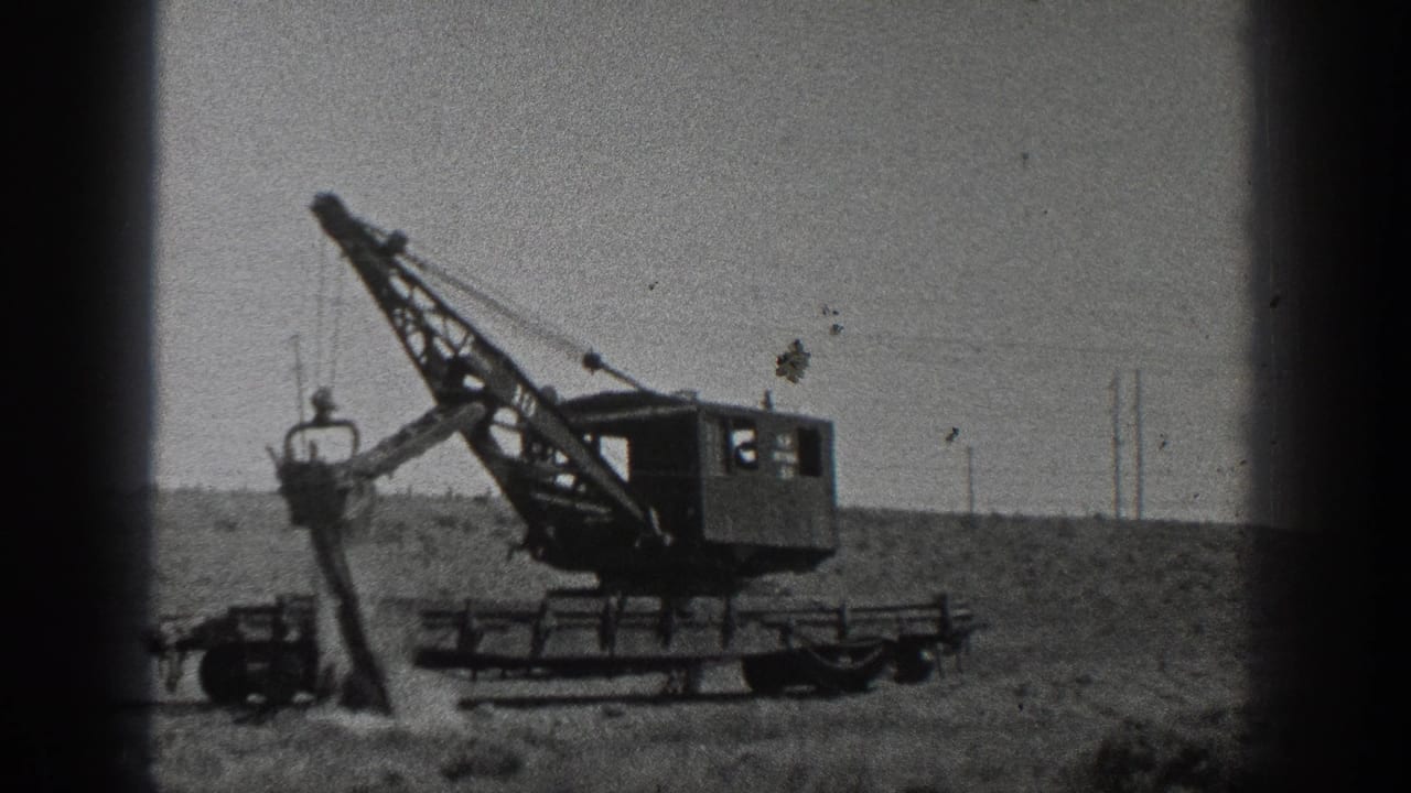 Crane boom raised with the bucket near a mound of soil in an open field.