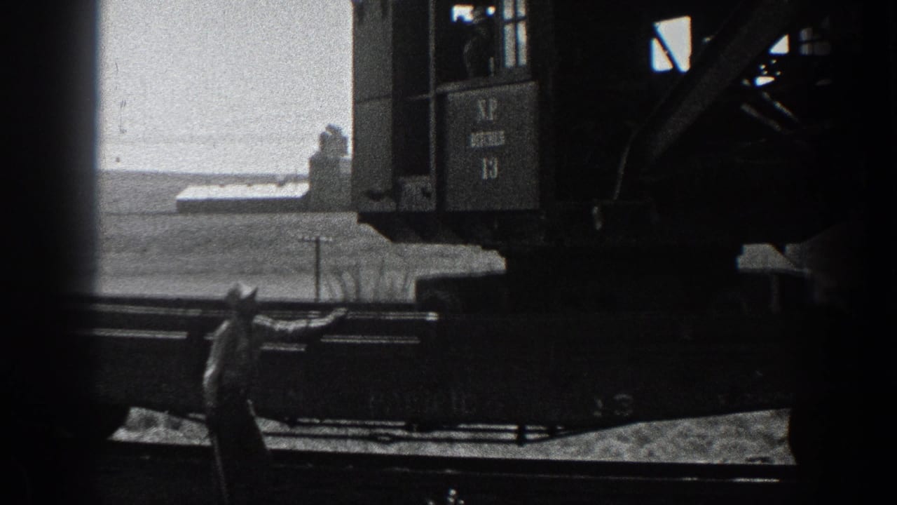 1936 California 8mm frame showing a rail-mounted industrial crane cab, a worker below and flat open terrain beyond.