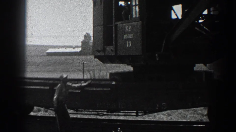 1936 California 8mm frame showing a rail-mounted industrial crane cab, a worker below and flat open terrain beyond.