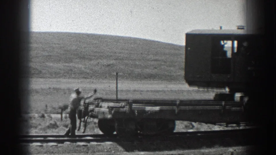 A worker remains beside the flat railcar as the rail crane sits partly out of frame against open California hills.