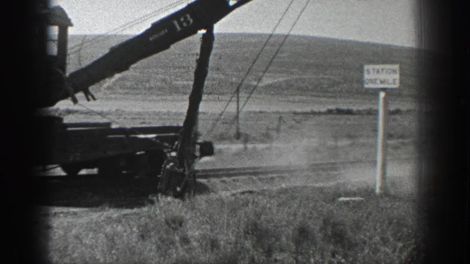 Black-and-white 1936 California frame of a crane marked 13 working near rail infrastructure, with dust, poles, hills, and a station-distance sign.