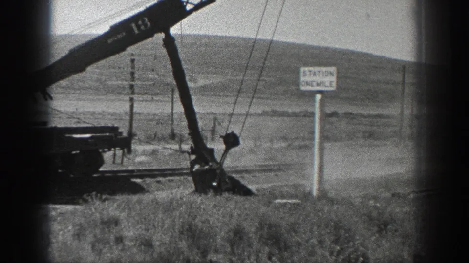 1936 California home-movie frame showing a rail-mounted crane lowering its arm near dusty ground beside utility poles and a STATION ONE MILE sign.