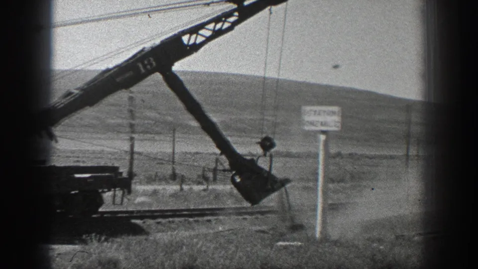 Black-and-white 8mm frame from 1936 California showing a crane marked 13, a hanging hook or load, dust, utility poles, and a STATION ONE MILE sign.