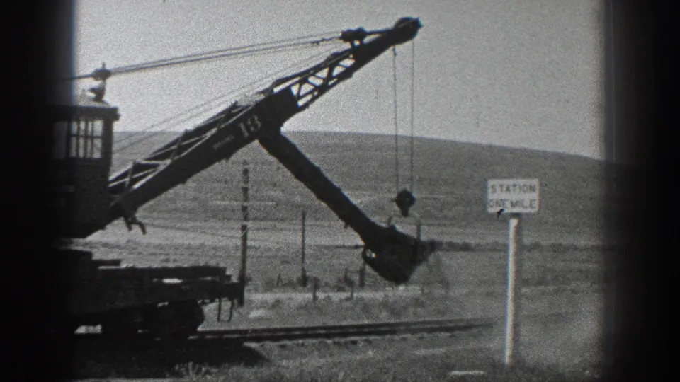 1936 California home-movie frame showing a rail-mounted crane marked 13 with a bucket near a station sign and low hills.