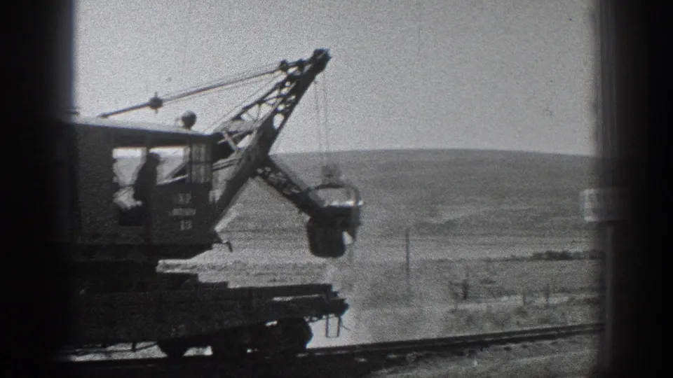 Home-movie frame from 1936 California showing the crane cab, boom, and bucket over rail tracks with hills beyond.