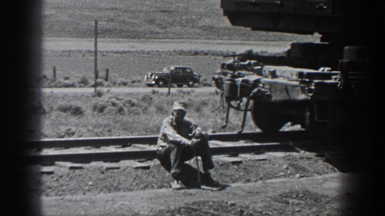 1936 California home-movie frame showing a man in work clothes seated by railroad tracks near rail equipment, with open rural land behind.