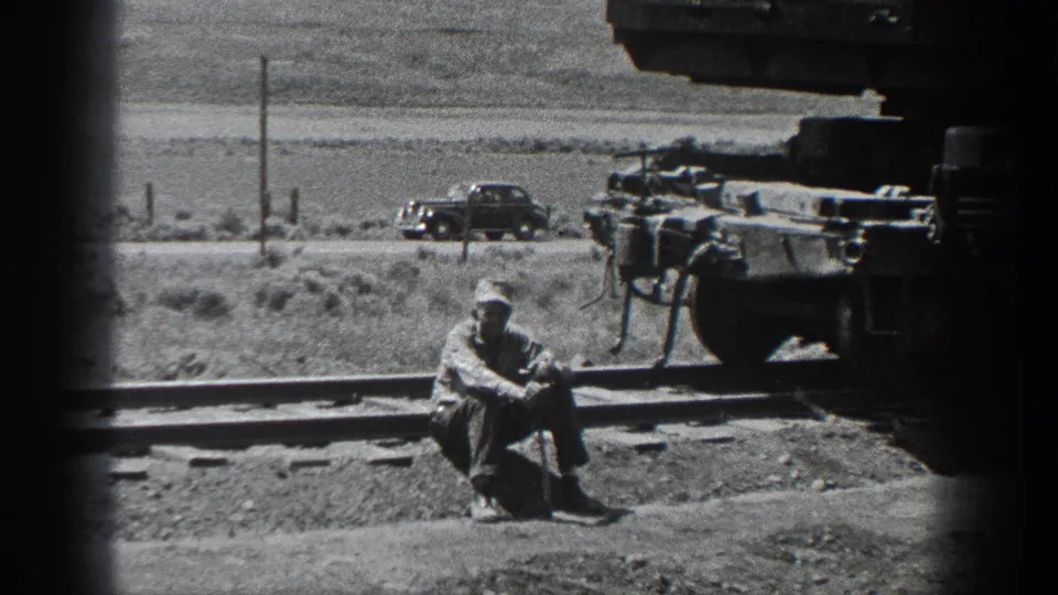 1936 California home-movie frame showing a man in work clothes seated by railroad tracks near rail equipment, with open rural land behind.