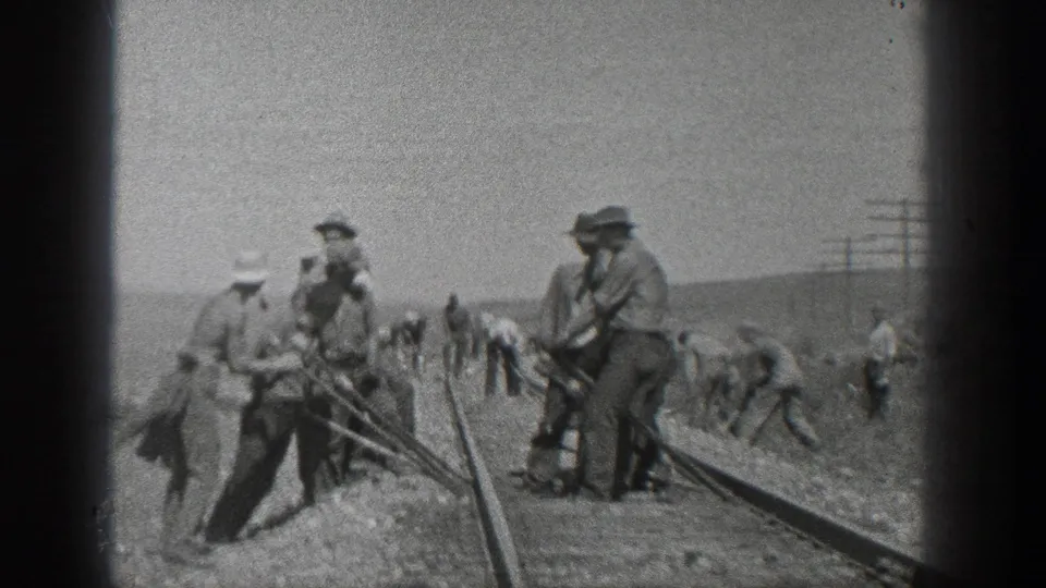 Men appear to maintain or inspect a curved railroad track in the California desert, with some standing near tools and others farther down the line.