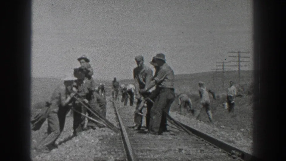 Railroad workers in period work clothes cluster beside the track and use tools, while additional men are visible along the rail bed in the open desert.