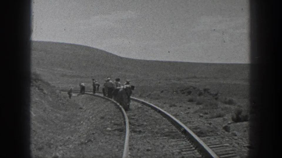 A wider frame shows the railroad track curving through desert hills as a small work crew stands along the rails under a bright open sky.