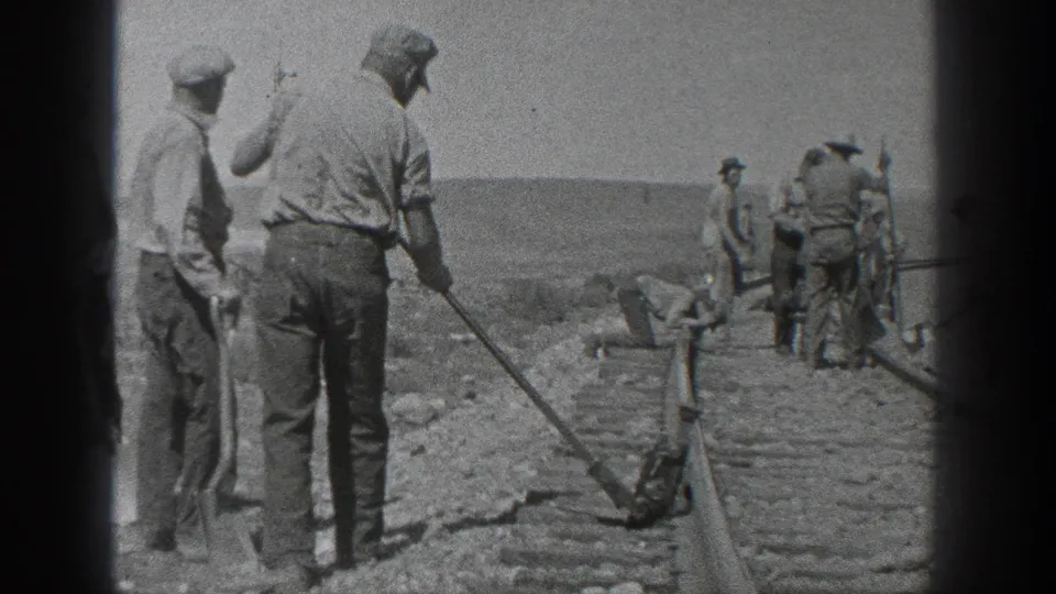 Black-and-white 1936 California 8mm frame showing adult men maintaining railroad tracks with shovels and hand tools in an open arid setting.