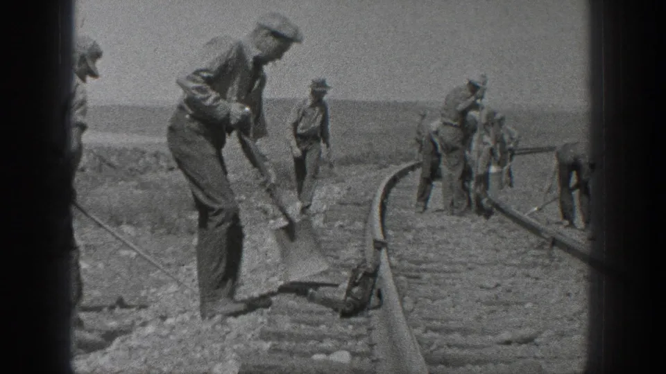 1936 California black-and-white home-movie frame showing a worker using a shovel beside railroad ties, with additional men and tools along the rail bed.