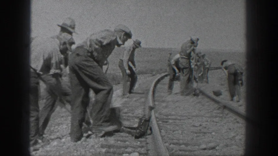 Black-and-white 8mm frame showing adult railroad workers beside tracks in California in 1936, with one foreground figure bending over a shovel and others spaced along the rail.