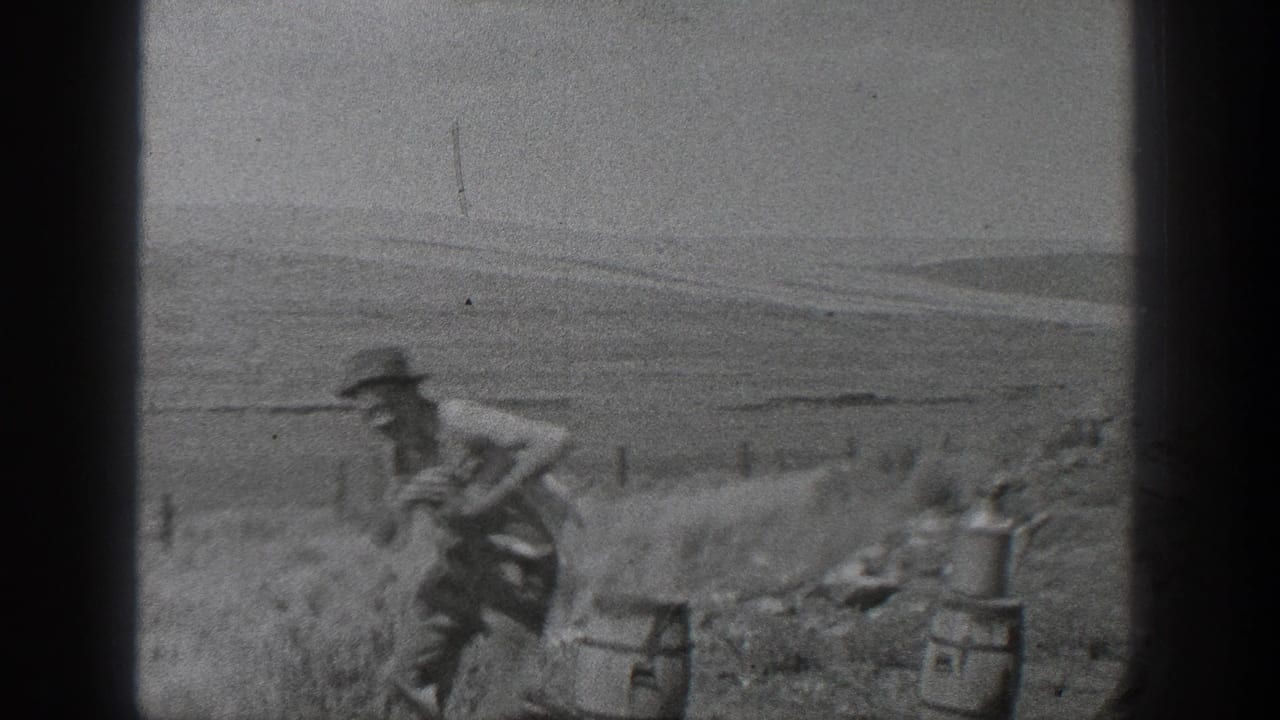 A worker in period clothing moves near rail materials and small equipment beside a rural track, with open land in the background.