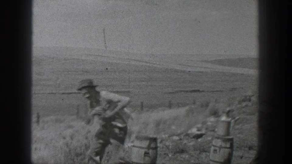 A worker in period clothing moves near rail materials and small equipment beside a rural track, with open land in the background.