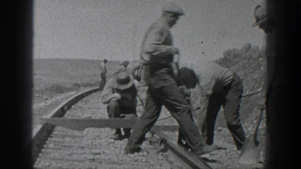 A group of men works at a railroad track in rural California, with one worker crossing the foreground in grainy black-and-white 8mm footage.
