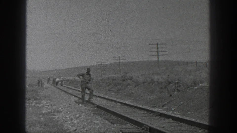 The rail line stretches through open terrain as one worker stands near the camera and the crew continues in the distance.