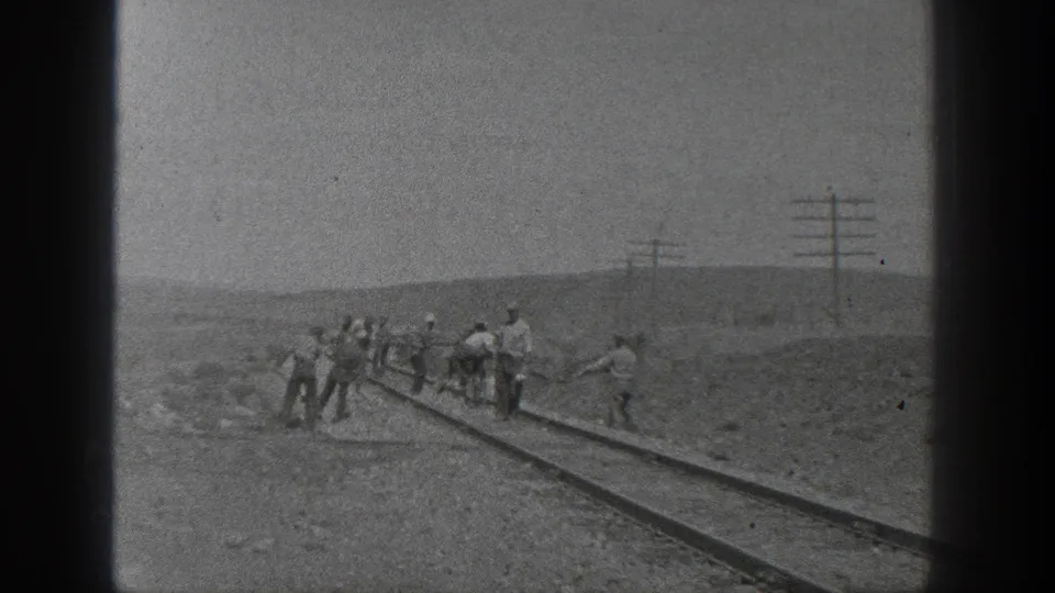 Black-and-white 8mm frame of railroad workers spread along a track in rural California, with utility poles beyond.