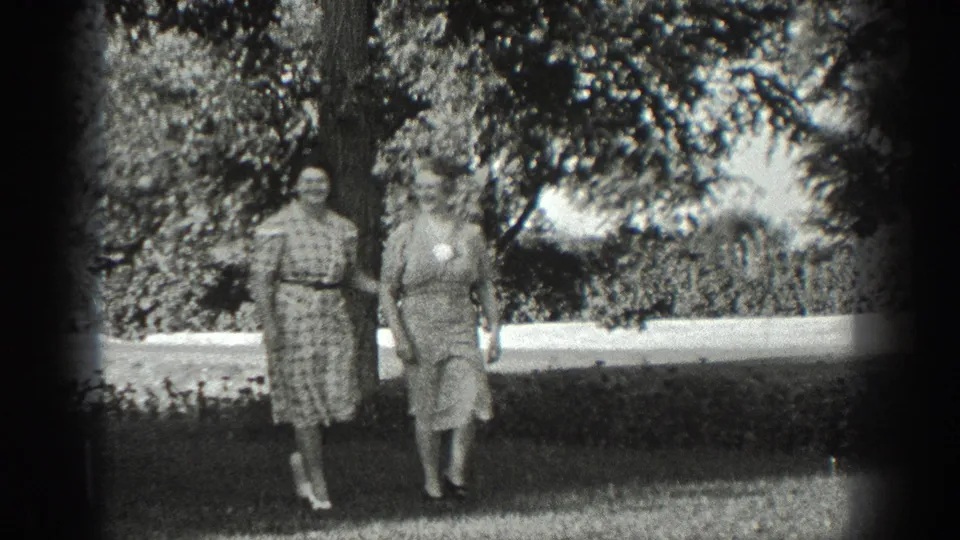 Two adult women in period dresses walk together through a California garden in 1936 home-movie footage.