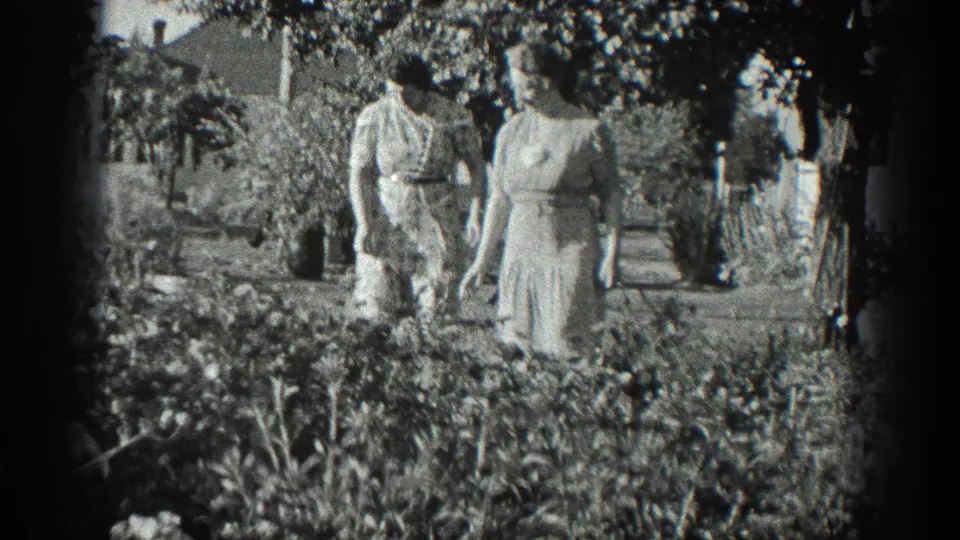 Two adult women in light dresses walk toward the camera along a planted garden path in the 1936 California footage.