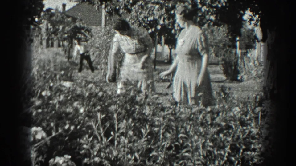 1936 California 8mm frame showing two older women in a garden, with a man working farther back near trees and low buildings.