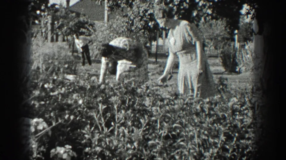Black-and-white 8mm frame from 1936 California showing one woman bending over a garden bed as another woman watches nearby.