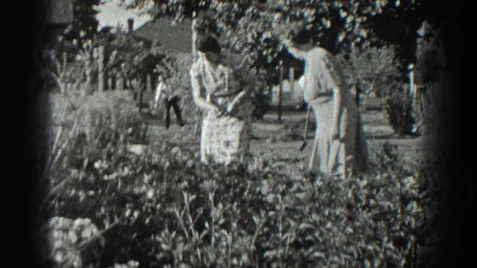 Black-and-white 8mm frame from 1936 California showing two women together among garden plants with a man working behind them.