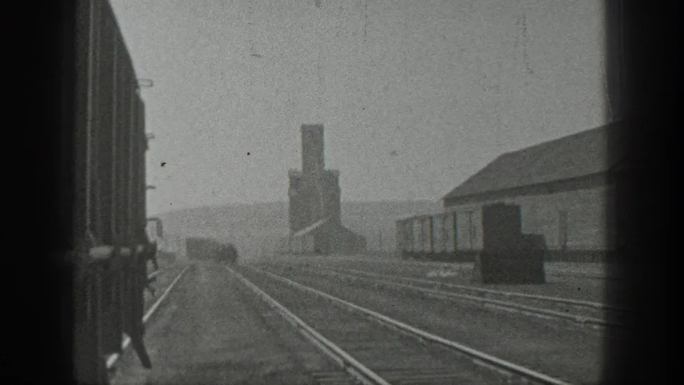 1936 California 8mm frame showing a quiet rail yard with empty tracks, low buildings, and a distant industrial structure.