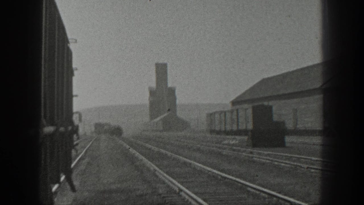 1936 California 8mm view from a moving train, showing parallel tracks, freight cars, a long building, and a distant smokestack.