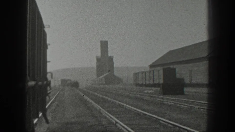 1936 California 8mm view from a moving train, showing parallel tracks, freight cars, a long building, and a distant smokestack.