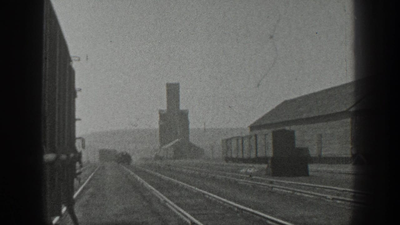 Black-and-white 8mm frame from California in 1936 showing rail tracks, freight cars, an industrial building, and a smokestack in the distance.
