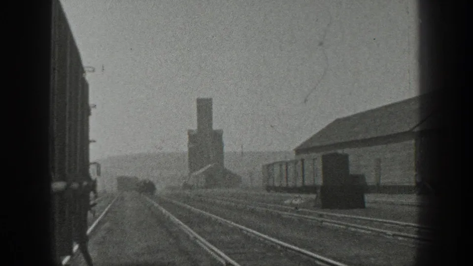 Black-and-white 8mm frame from California in 1936 showing rail tracks, freight cars, an industrial building, and a smokestack in the distance.