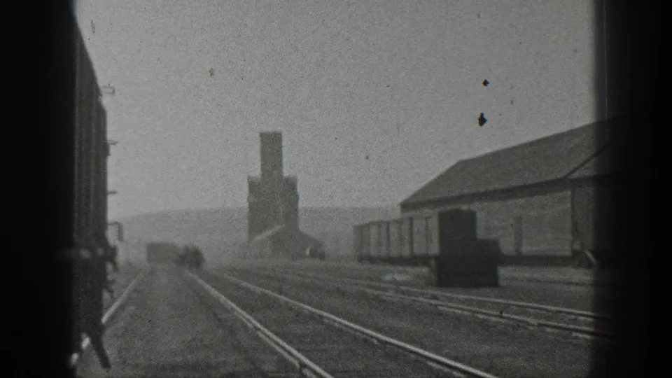 Grainy 1936 California train-view frame with parallel tracks, yard structures, freight cars, and a tall smokestack under a hazy sky.