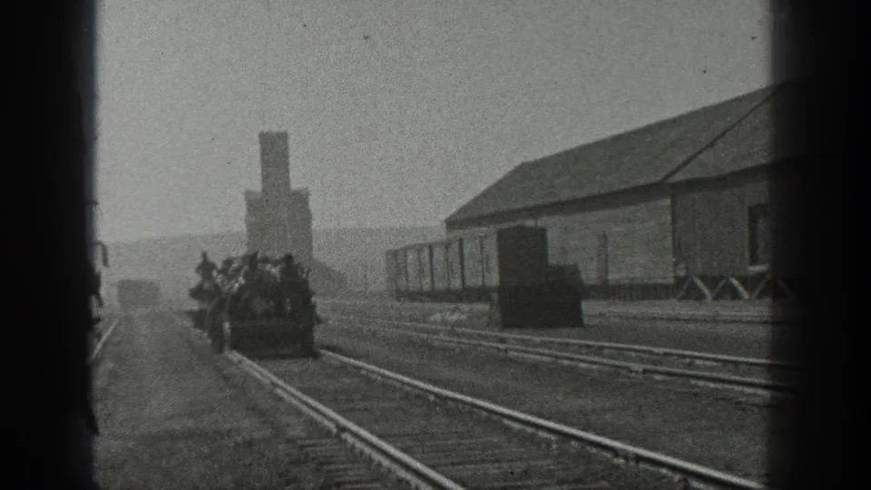 1936 California 8mm frame showing several distant people riding an open rail car along tracks beside industrial buildings and freight cars.