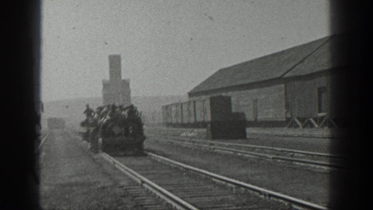 Crowded open rail car approaches along tracks beside a long industrial building.